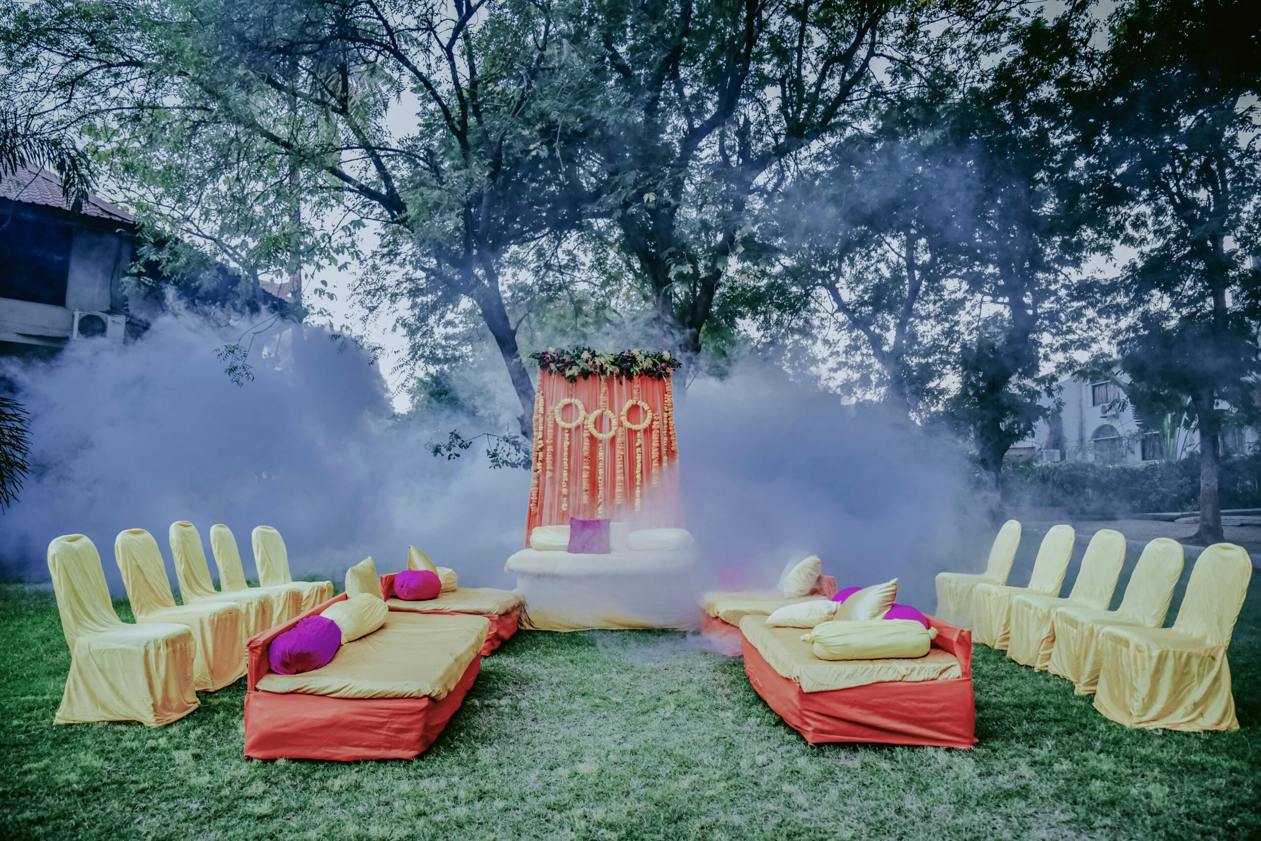 Colorful mehendi ceremony setup with smoke effects, featuring yellow and red decor in an outdoor garden setting.